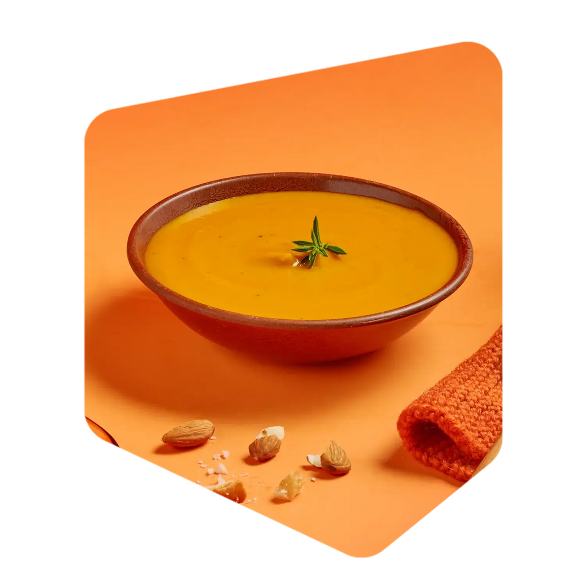 Bol de velouté de carotte Pocket Soupe garni de romarin, avec des amandes et un linge de cuisine orange